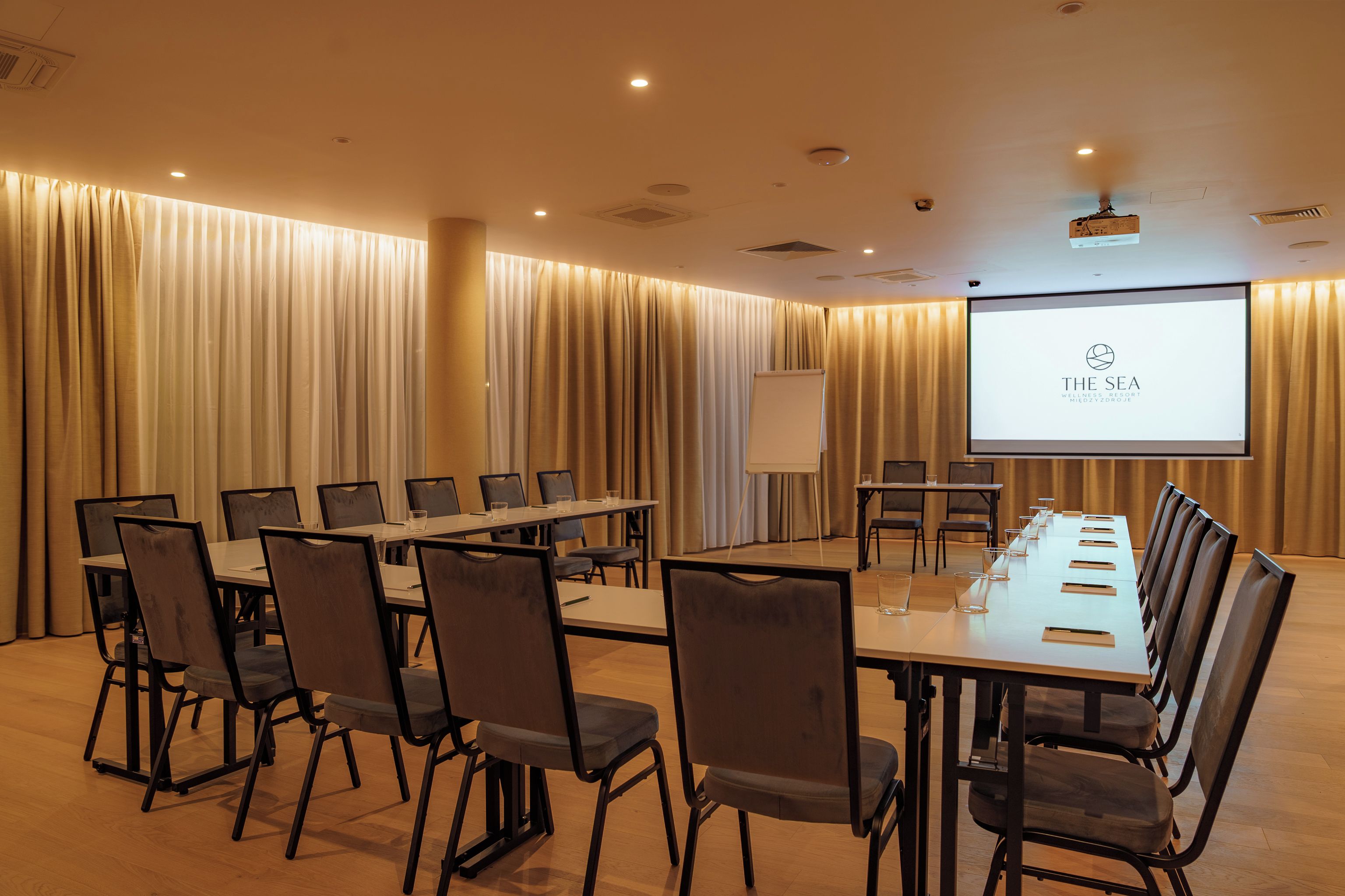 A conference room with a long table, chairs, a projector screen, and curtains.