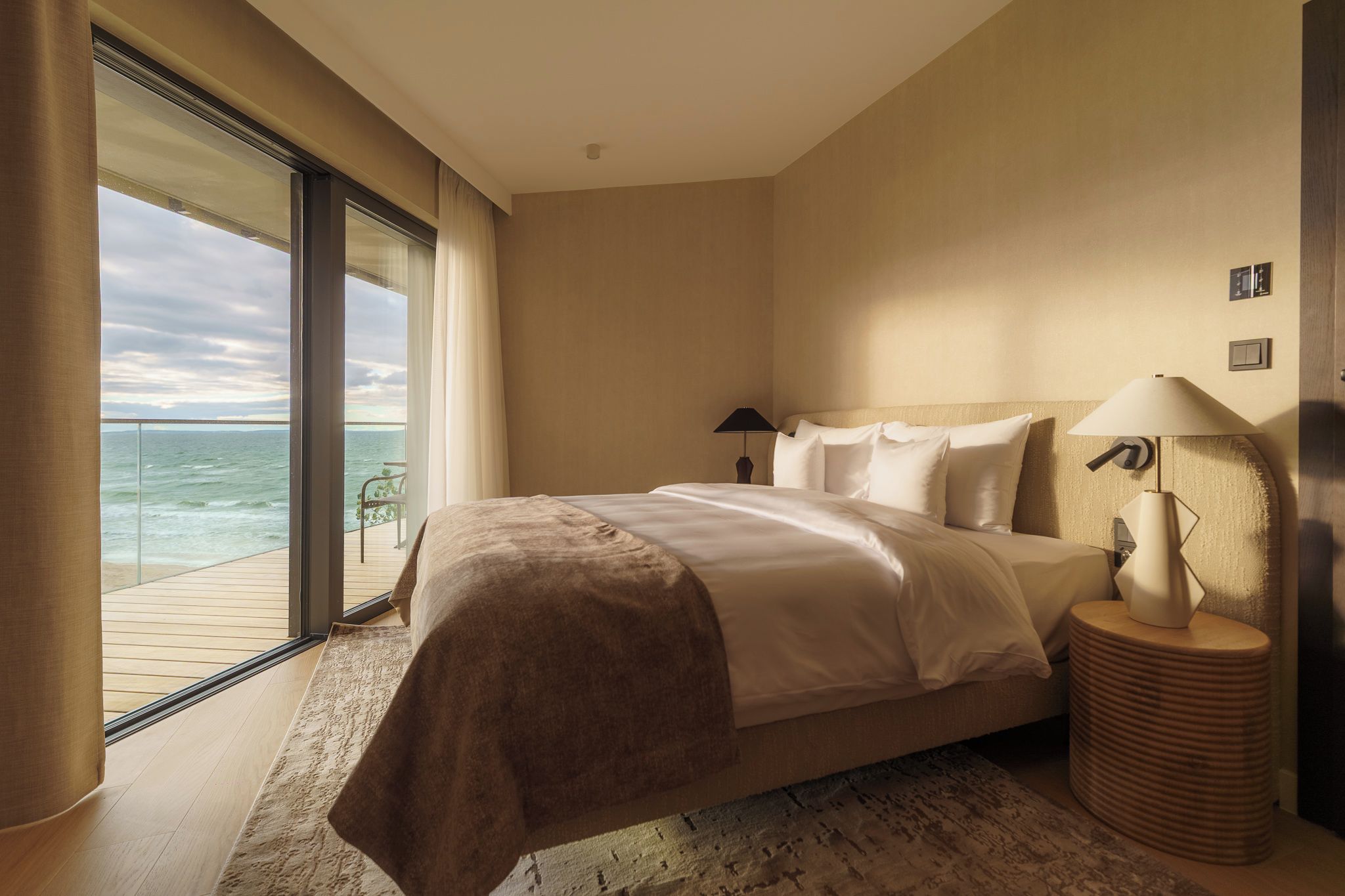 Ein Schlafzimmer mit einem großen Bett, einem Nachttisch, einer Lampe und Blick auf das Meer durch eine Schiebetür.
