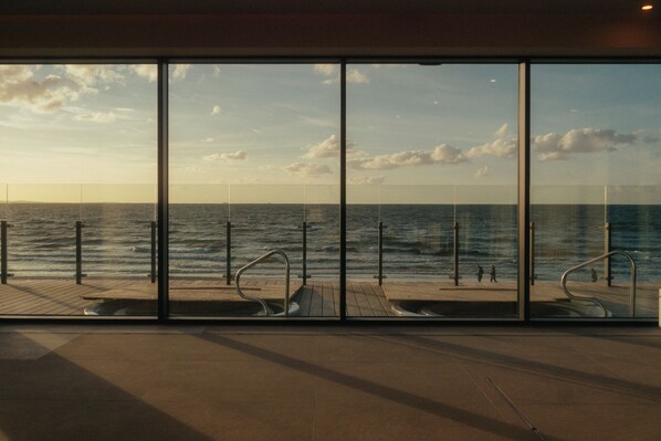 A view through large windows of a wooden deck with metal railings overlooking a body of water.
