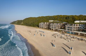 A beachfront hotel with multiple levels, a sandy beach, and a forested hillside in the background.