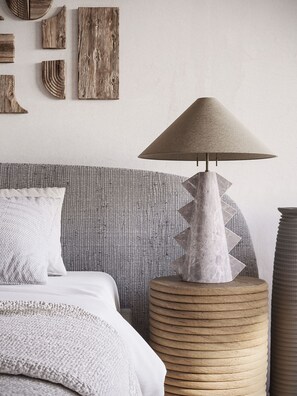 A bed with a patterned headboard, a round woven side table, and a conical lamp.
