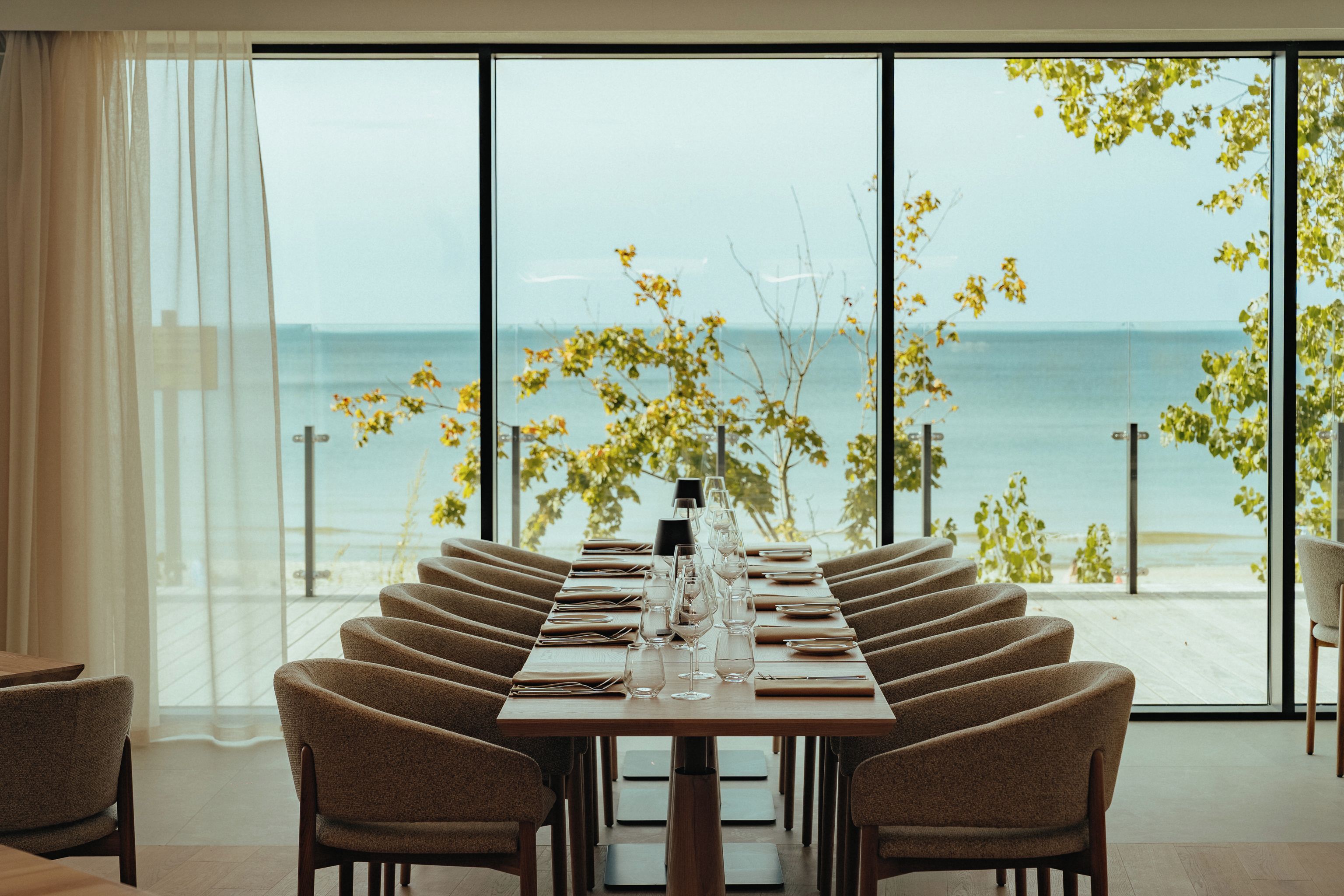 Ein Esszimmer mit gedecktem Tisch, Stühlen, großen Fenstern mit Meerblick und einem Vorhang auf der linken Seite.