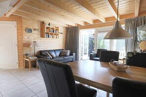 Ein gemütliches Zimmer mit Holzbalkendecke, einer Couch, einem Esstisch mit Stühlen, einem Bücherregal und einem großen Fenster mit Vorhängen.