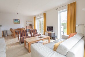Ein Wohnzimmer mit Sofa, Sessel, Couchtisch und Fernseher. Es gibt einen Esstisch mit Stühlen und ein Bücherregal.