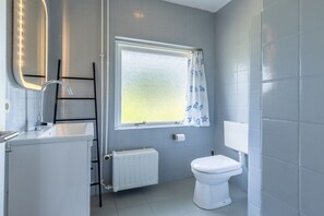 A bathroom with a white sink, a mirror with lights, a toilet, a window with a curtain, and a radiator. - Domburgseweg 52 Apartment (Oostkapelle)