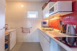 A compact kitchen with white cabinets, a microwave, a kettle, and a small window. - Domburgseweg 52 Apartment (Oostkapelle)