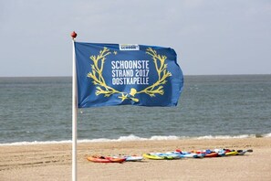 A flag with 'Schoonste Strand 2017 Oostkapelle' text and a yellow emblem, with kayaks lined up on the beach. - Domburgseweg 52 Apartment (Oostkapelle)