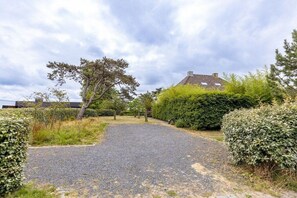 A gravel driveway leading to a house with a sloped roof, surrounded by dense shrubbery and a large tree. - Domburgseweg 52 Apartment (Oostkapelle)