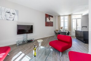 Ein modernes Wohnzimmer mit Fernseher, roten Sesseln, einem Glastisch und einem Essbereich mit Tisch und Stühlen.