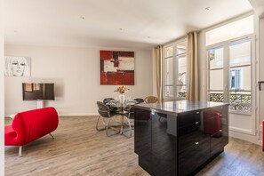 Un salon moderne comprenant un coin repas, un fauteuil rouge, un meuble noir et une grande fenêtre avec des rideaux.