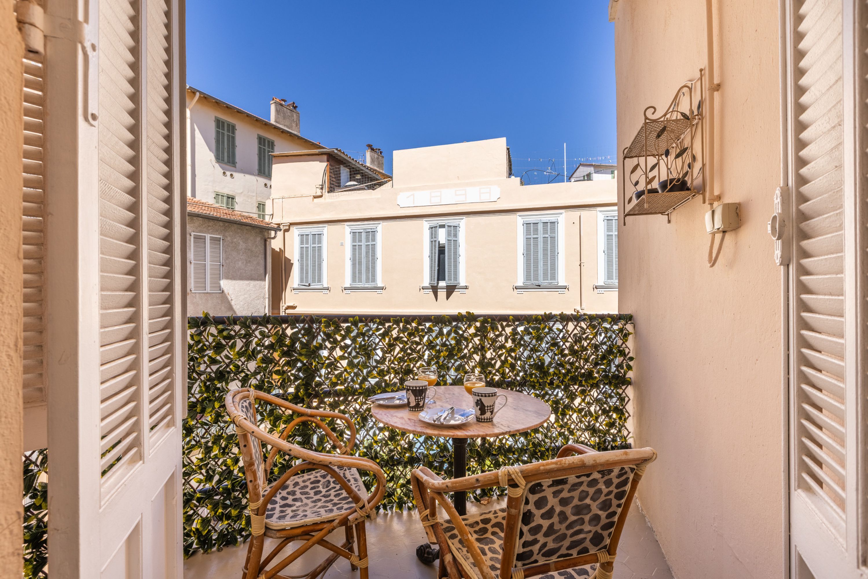 A balcony with two wicker chairs and a small round table, overlooking a row of buildings with shutters.