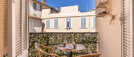 A balcony with two wicker chairs and a small round table, overlooking a row of buildings with shutters.