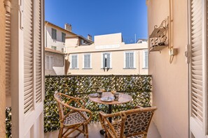 Un balcon avec deux fauteuils en osier et une petite table ronde, donnant sur une rangée d’immeubles aux volets fermés.
