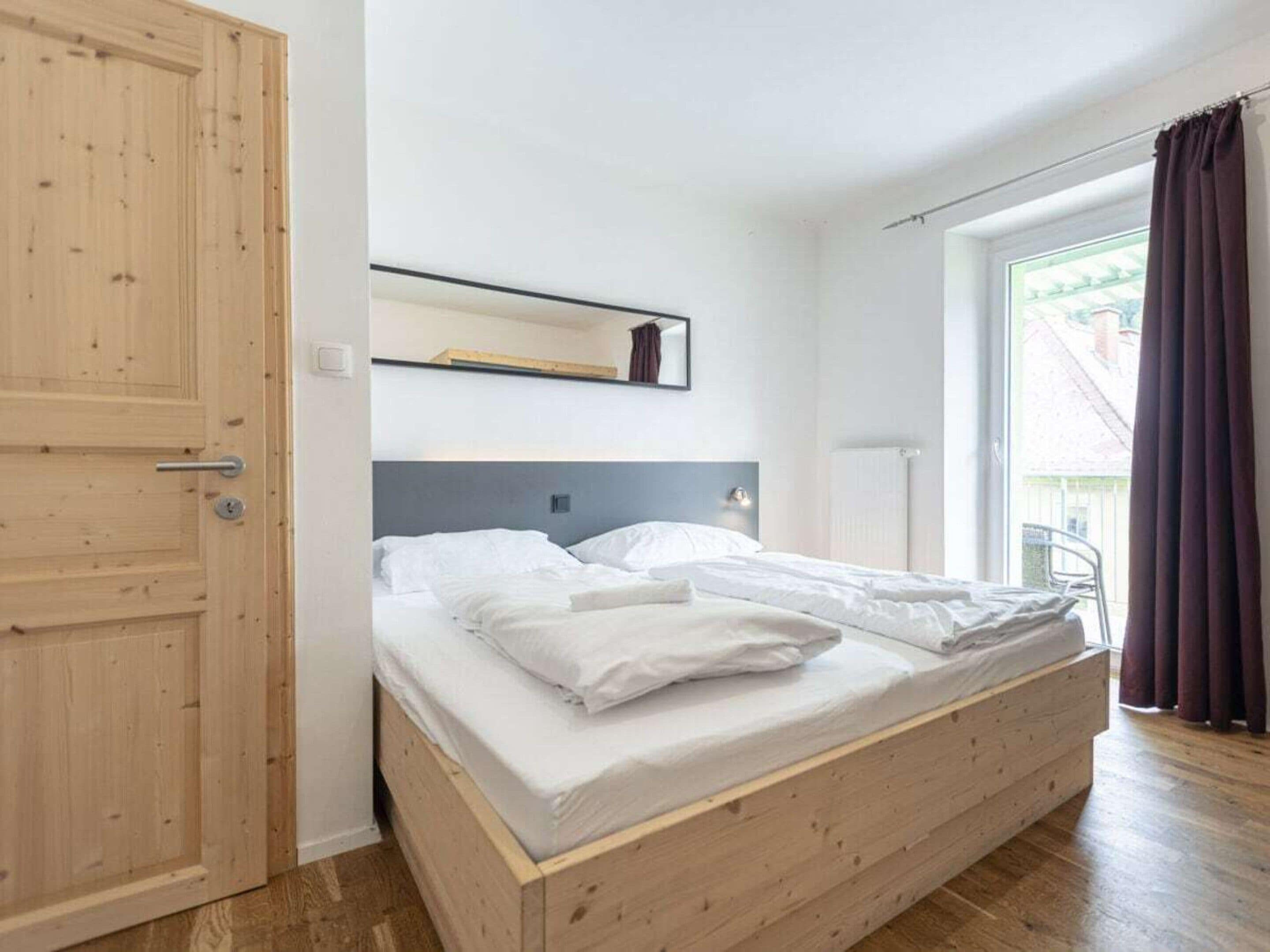 A single bed with a wooden frame and white bedding in a room with a wooden door and a window with curtains.