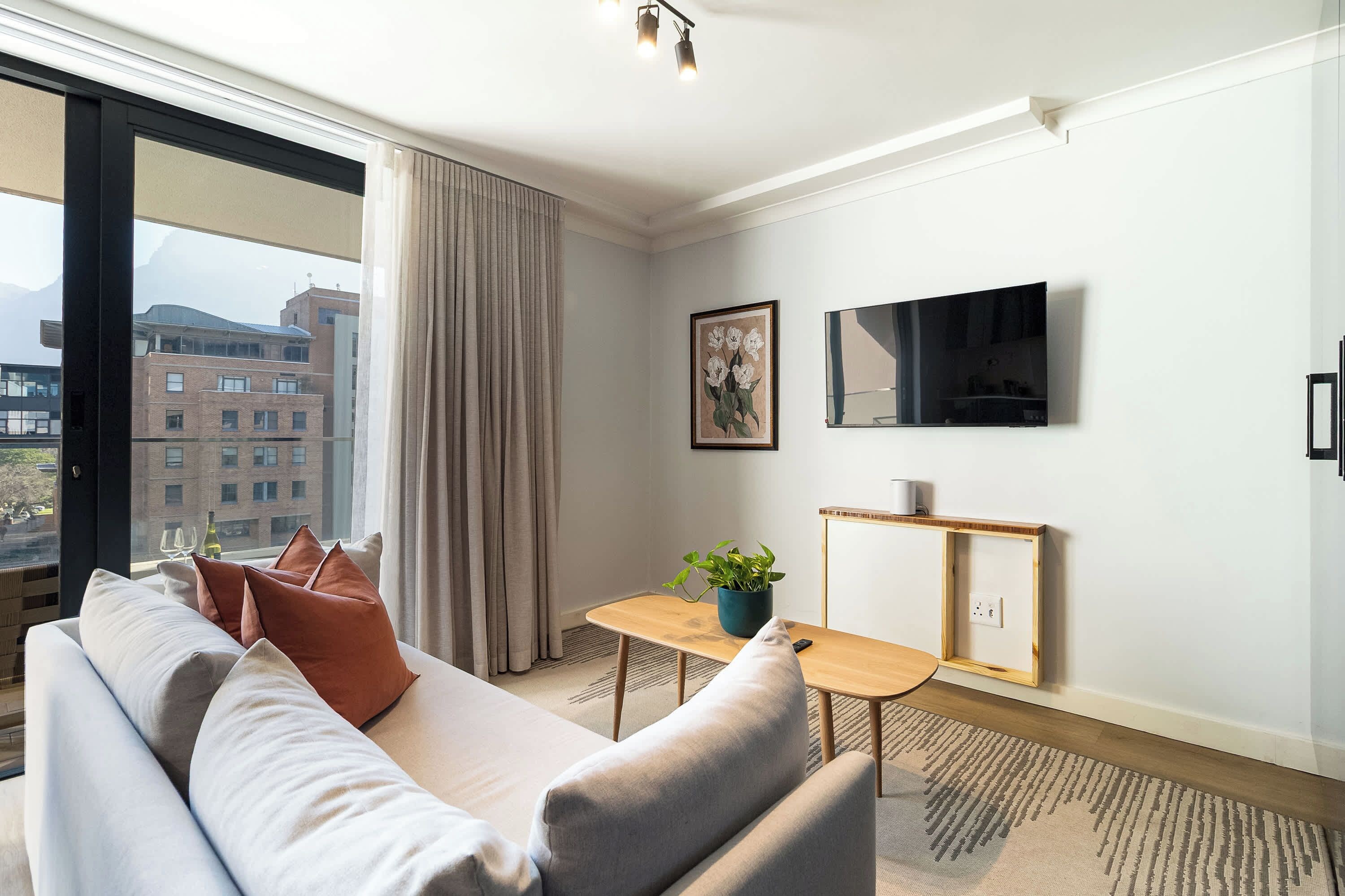 A modern living room with a sofa, a small table with a plant, a mounted TV, and a window with a view of buildings.