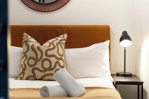 Ein ordentlich gemachtes Bett mit einem gemusterten Kopfkissen und einem zusammengerollten Handtuch, eine Nachttischlampe und ein Wandspiegel.