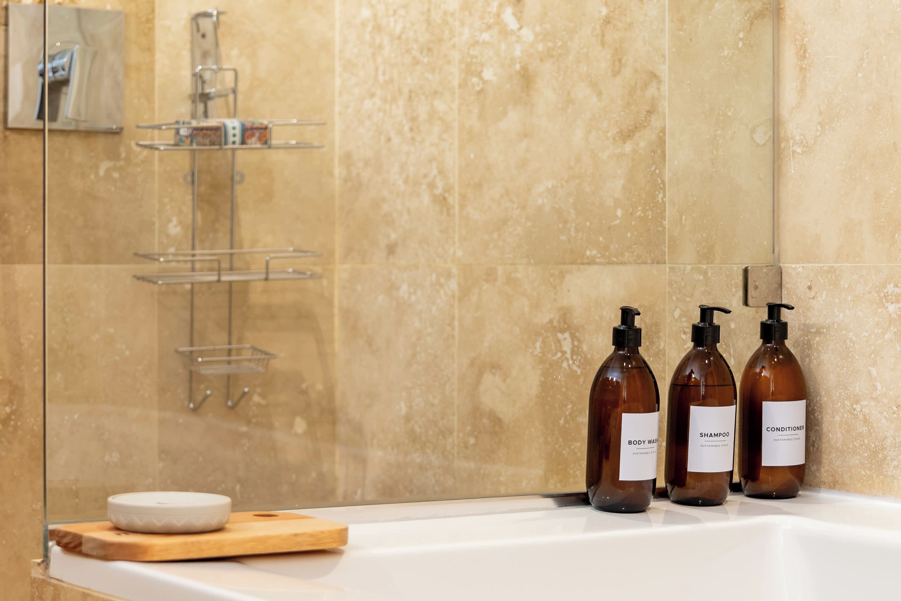 A bathroom with a glass shower enclosure, a wooden soap dish, and three bottles of body wash and conditioner.