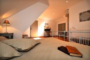 Une chambre avec un plafond en pente, un lit recouvert d’une courtepointe, une lampe de chevet, un bureau avec une chaise et une vue sur un coin cuisine.