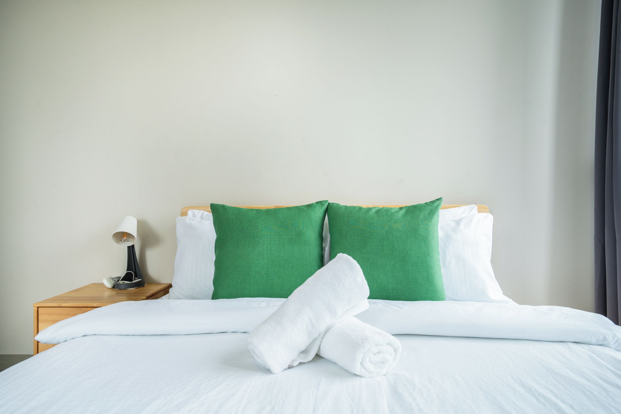 A neatly made bed with white linens, two green pillows, and a rolled white towel.