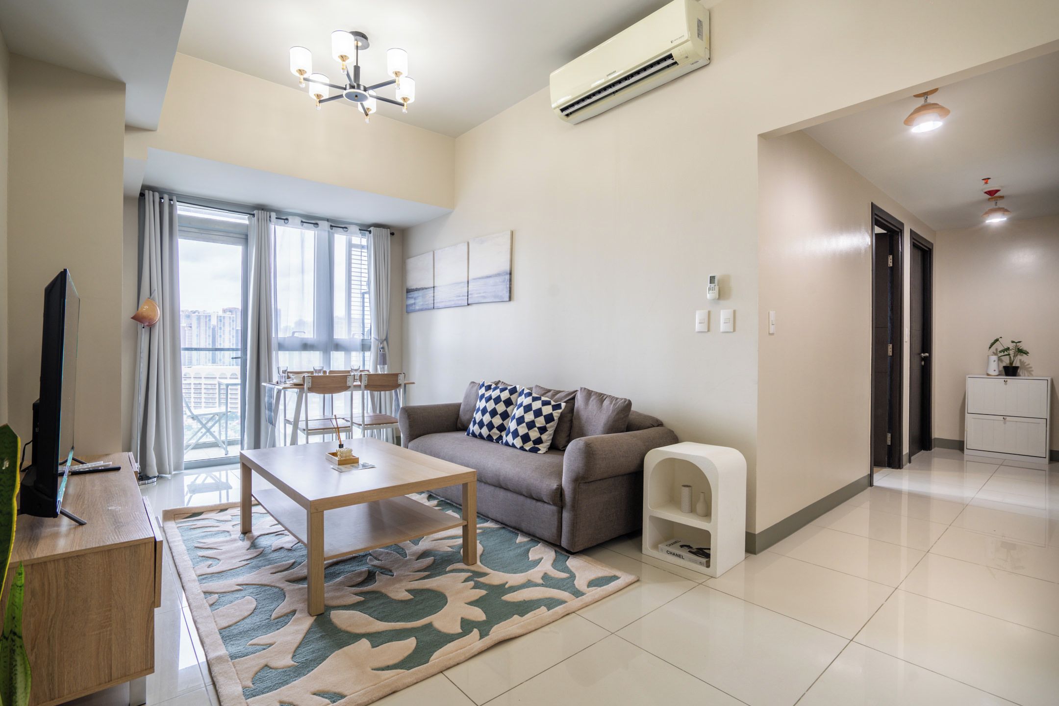 A modern living room with a sofa, coffee table, and a TV. There is a large window with curtains, an air conditioning unit, and a chandelier.