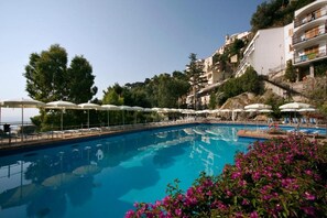 A swimming pool with sun umbrellas and lounge chairs, surrounded by buildings and greenery.