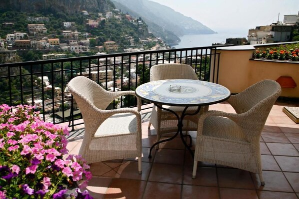 A balcony with wicker chairs and a table, overlooking a coastal town and the sea.