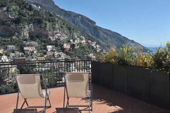 A balcony with two chairs, overlooking a coastal town with buildings on a hillside.