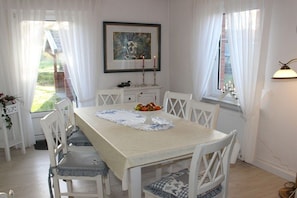A dining area with a table set for four, white chairs, a white tablecloth, a fruit bowl, and a framed painting on the wall. - Holiday home with 2 bedrooms (Dornumersiel)