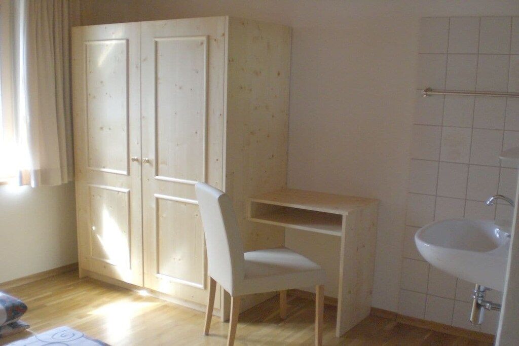 Une pièce comprenant une armoire en bois, une chaise blanche, un petit bureau en bois et un lavabo blanc.