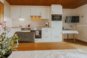 A compact kitchen with white cabinets, a stove, and a microwave. There's a small table with a white tablecloth and a clock on the wall. - Holiday apartment with 2 bedrooms (Gurtis)