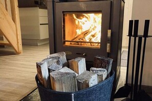 A wood-burning stove with visible flames, a storage bin with cut logs, and a black metal stand with candles. - Ski lodge in Ebene Reichenau with sauna (Ebene Reichenau)