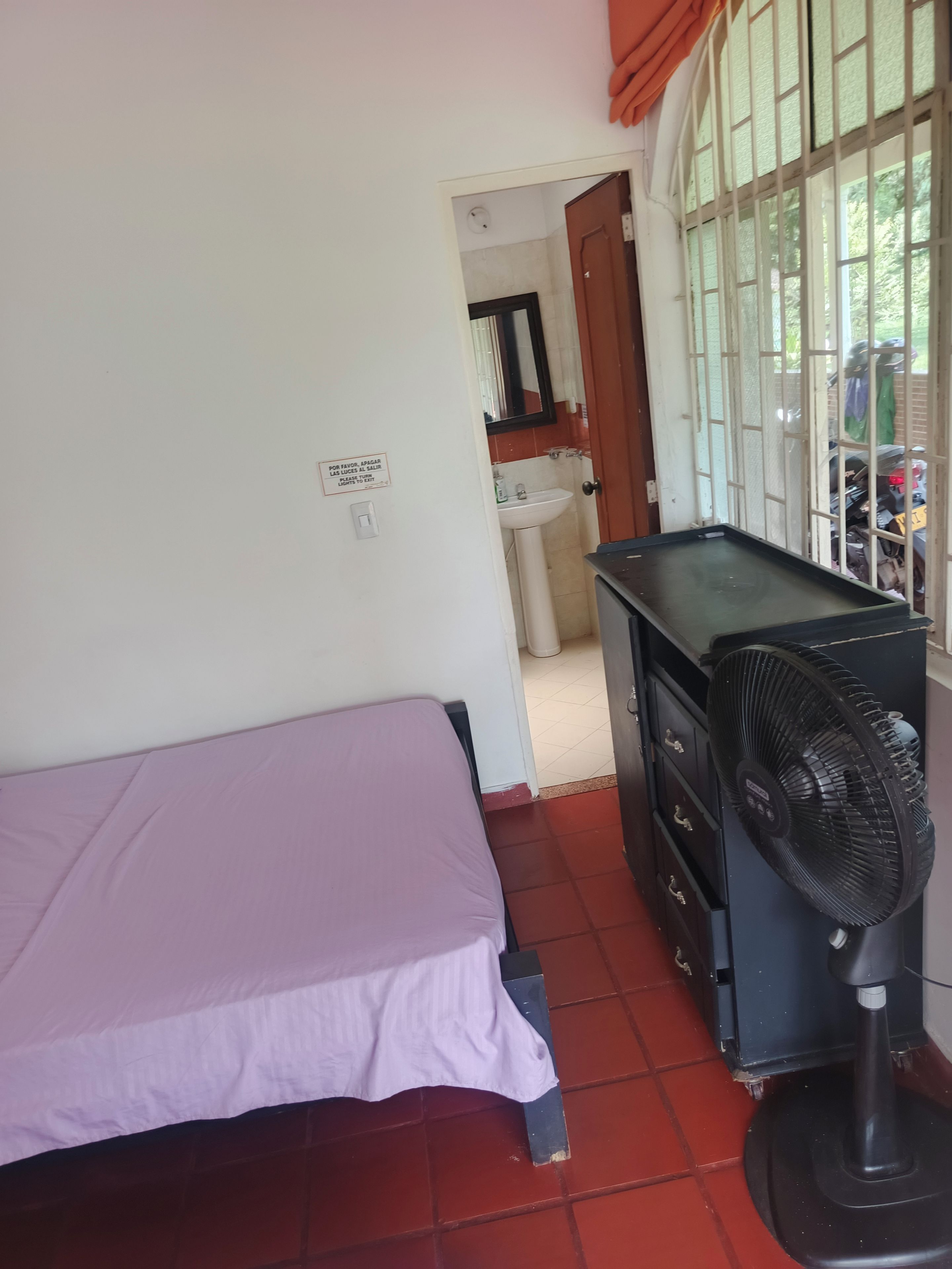 Quarto com cama, escrivaninha, ventoinha e janela com grades.