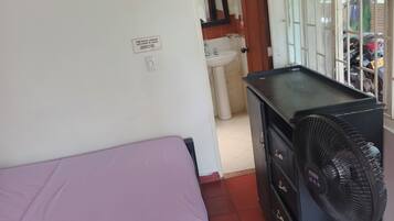 Quarto com cama, escrivaninha, ventoinha e janela com grades.