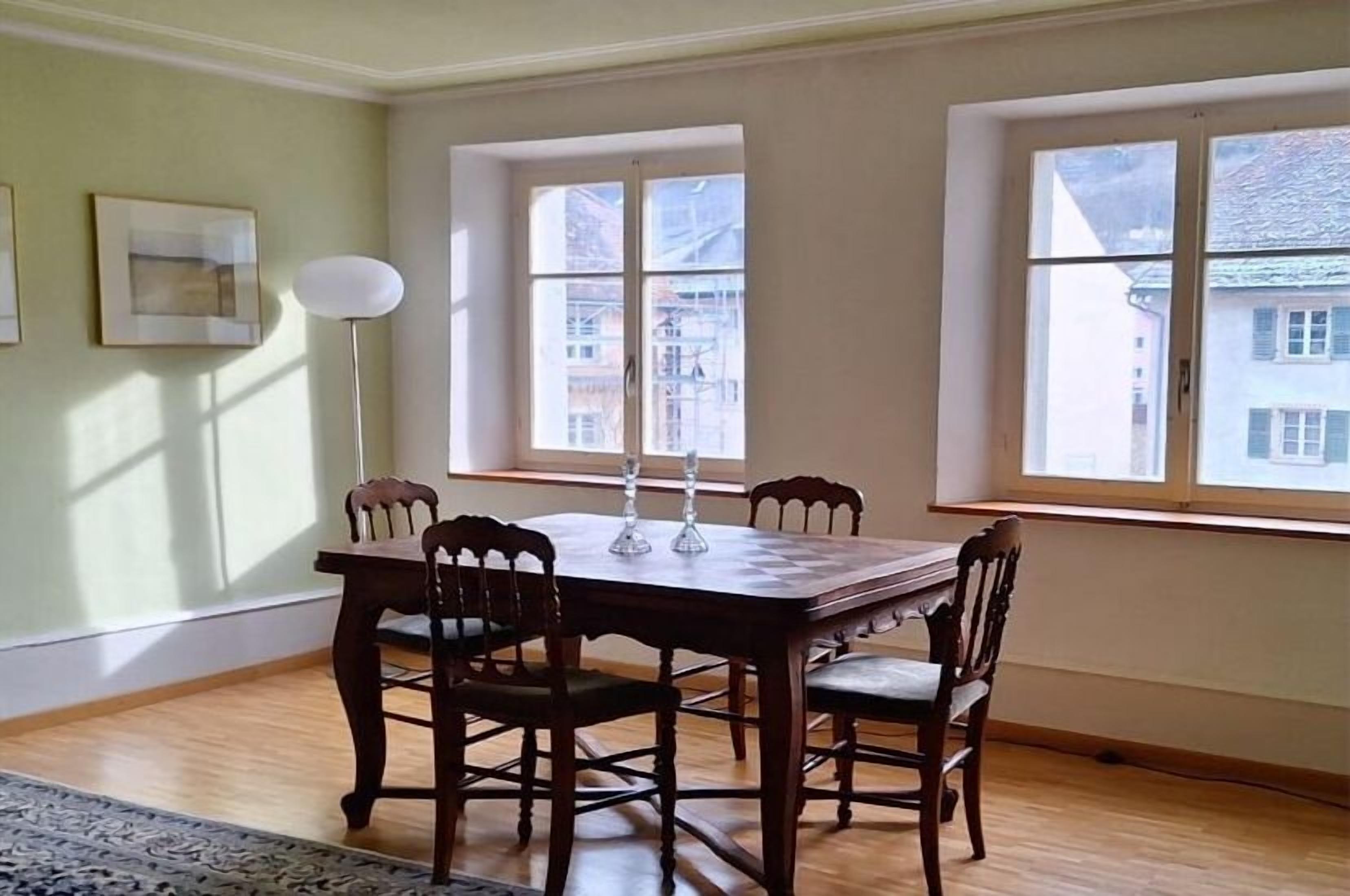 Un comedor con mesa y sillas de madera, ventanas amplias y una lámpara de pie.