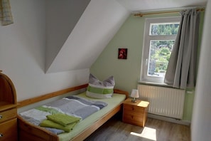 Een kleine kamer met een enkel bed, een schuine kap, een raam met gordijnen, een nachtkastje met een lamp en een radiator.
