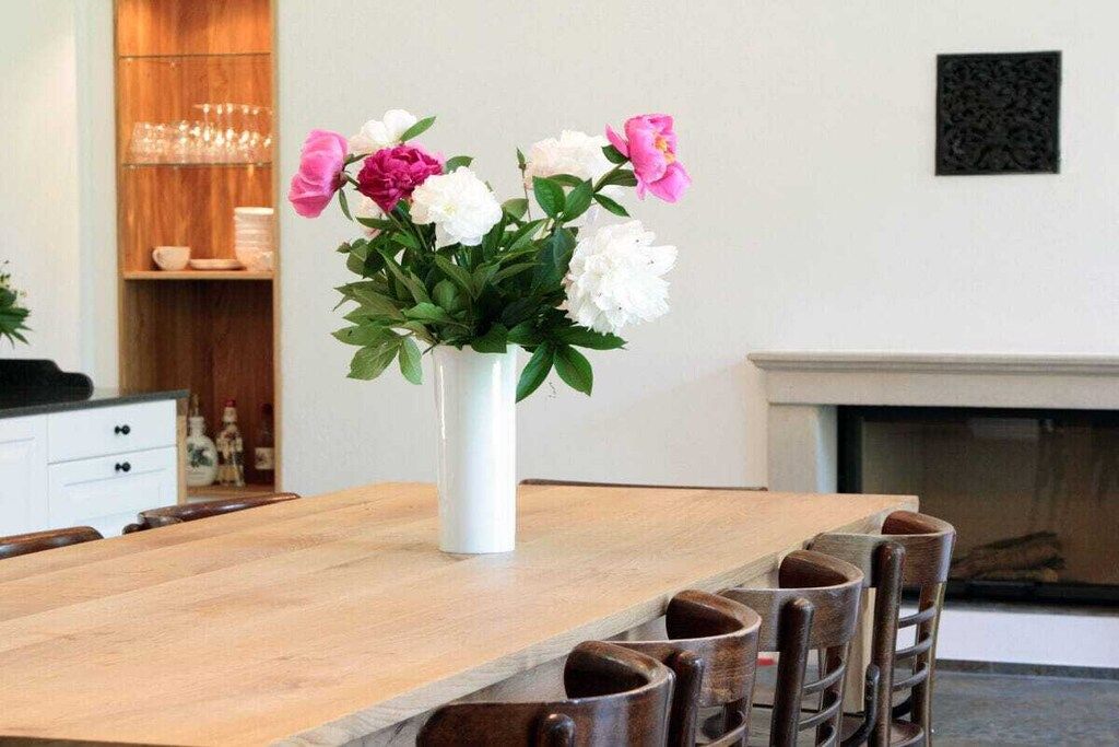 A dining table with a white vase of flowers, wooden chairs, and a fireplace in the background.