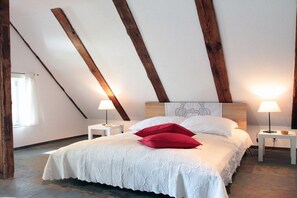 Ein Schlafzimmer mit schrägen Wänden, einem Bett mit weißer Bettwäsche und roten Kissen, zwei Nachttischen mit Lampen und einem Fenster mit Vorhängen.