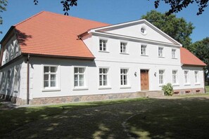 Ein zweistöckiges weißes Gebäude mit rotem Dach, einem zentralen Eingang mit einer Holztür und einem Steinfundament.