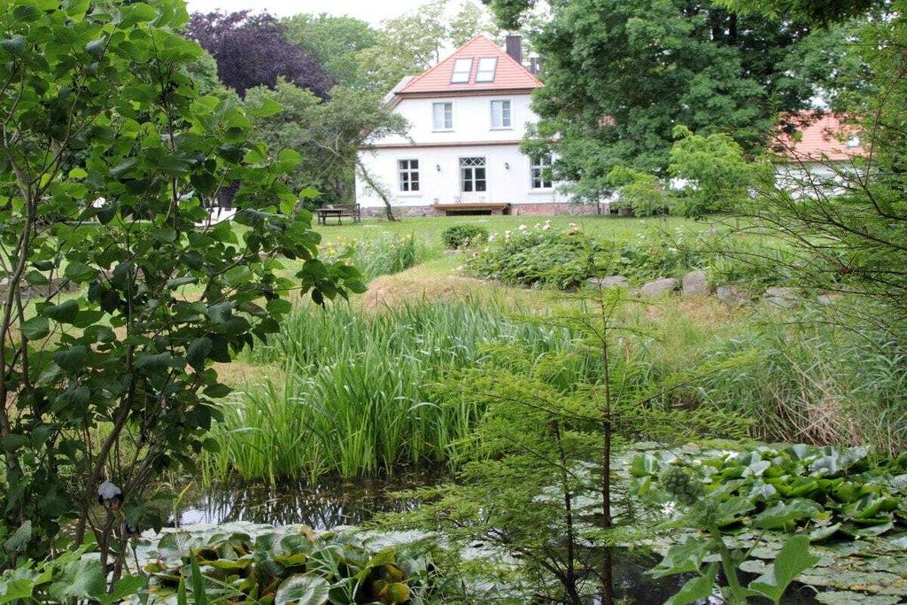 A two-story house with a red roof, surrounded by lush greenery and a pond.