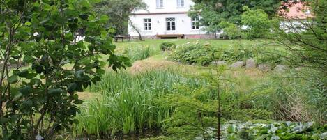 A two-story house with a red roof, surrounded by lush greenery and a pond.