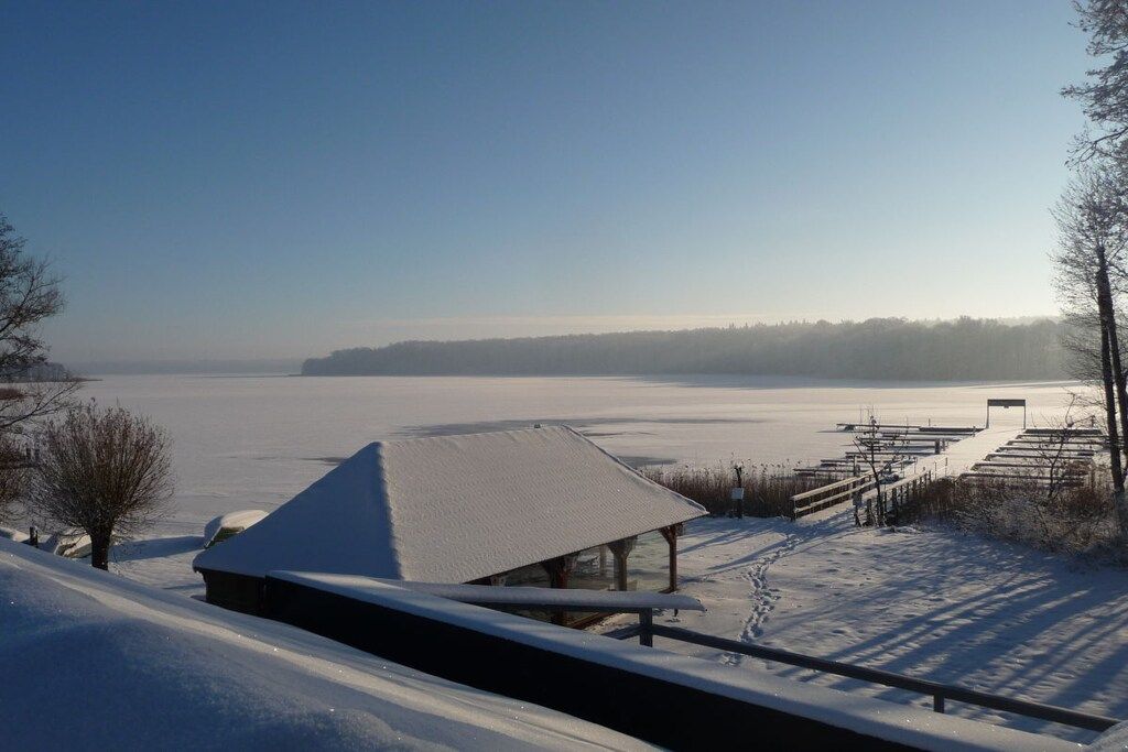 Paesaggio innevato con una struttura in legno, un lago ghiacciato e cielo sereno.