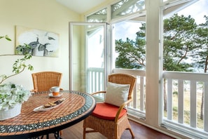 A room with a wicker chair, a table with a tiled top, a white flower arrangement, and a view of trees through a glass door. - Holiday apartment with 1 bedroom (Ostseebad Binz)