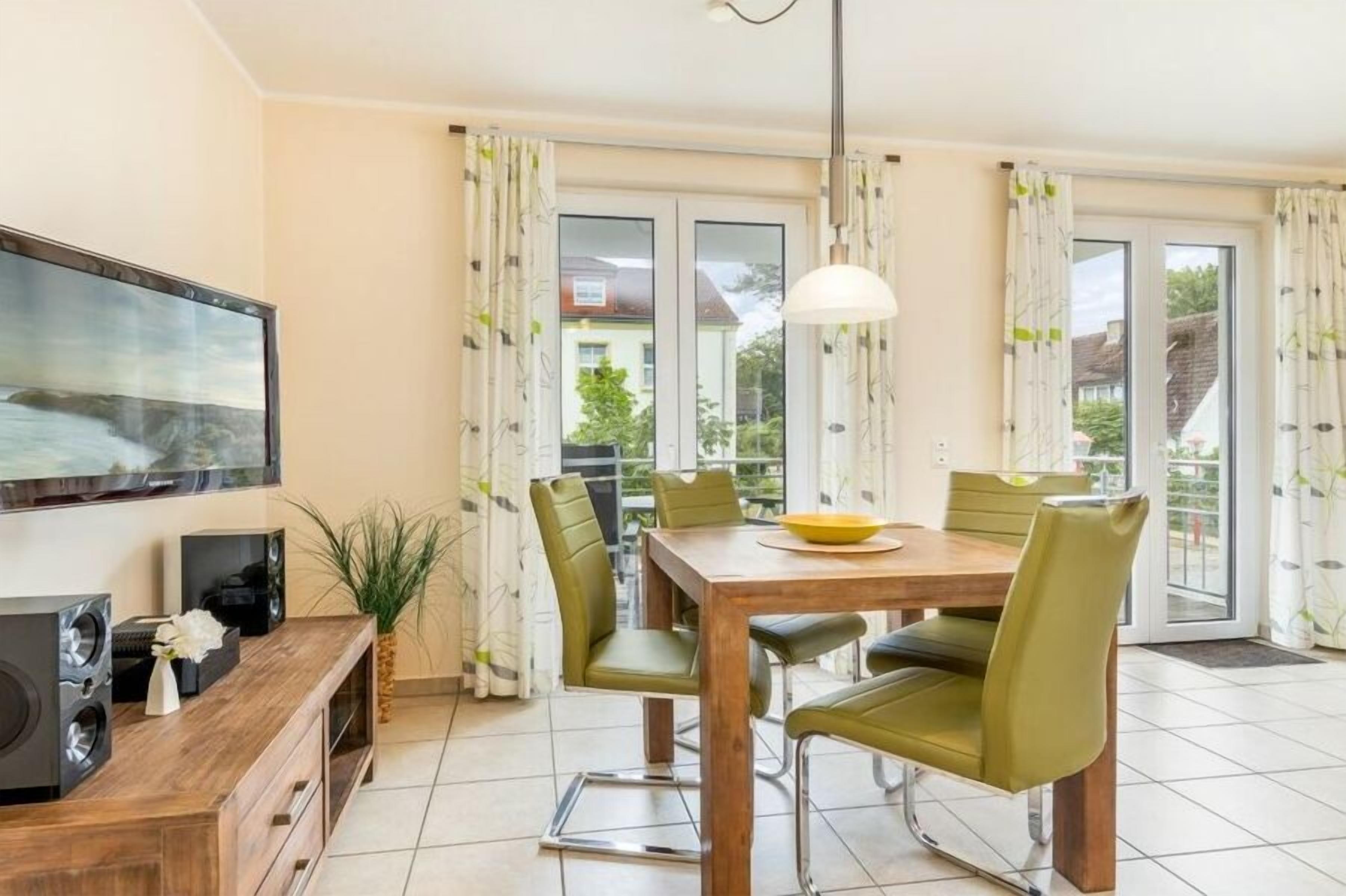 Ein Essbereich mit Holztisch, grünen Stühlen, einem Fernseher und einem großen Fenster mit Vorhängen.