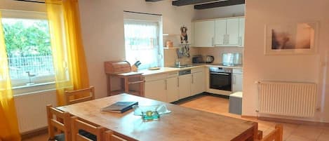 A dining area with a wooden table and chairs, a kitchen with white cabinets, and a window with yellow curtains.