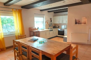 A dining area with a wooden table and chairs, a kitchen with white cabinets, and a window with yellow curtains. - Holiday home with 2 bedrooms (Göhren-Lebbin)