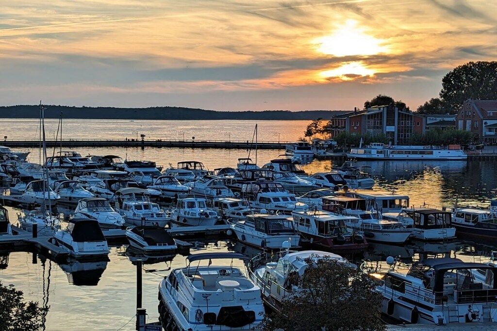 A marina with multiple boats docked, a sunset in the background, and buildings along the waterfront.