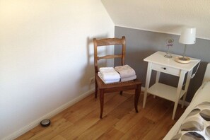 A wooden chair with folded towels, a small white nightstand with a lamp, and a white bed with a patterned cover. - Holiday apartment with 2 bedrooms (Kappeln)