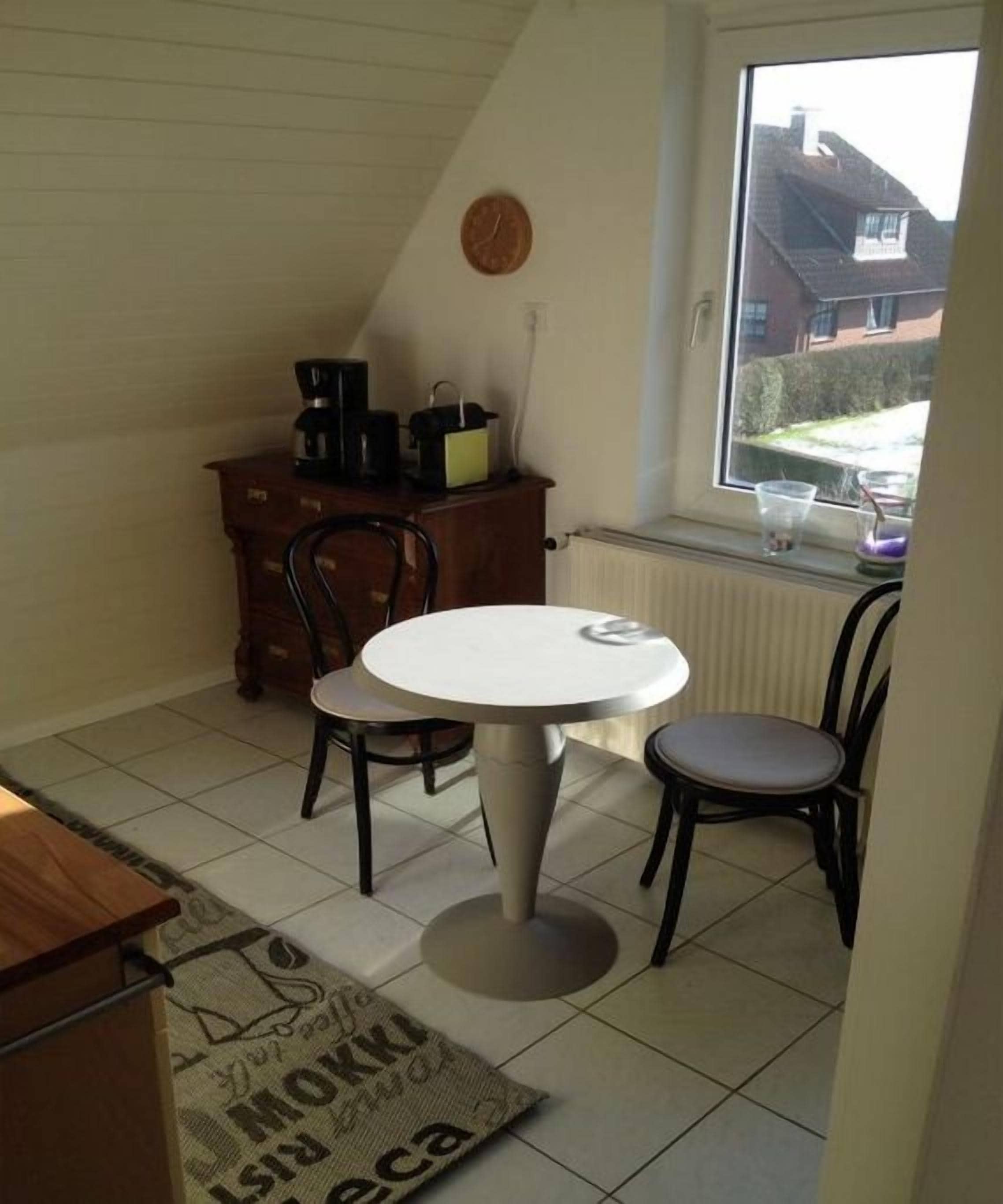 Ein kleiner Essbereich mit einem runden Tisch, vier Stühlen, einer Kaffeemaschine und einem Fenster mit Blick auf ein Haus.