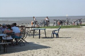 Ein Essbereich direkt am Strand mit Tischen und Stühlen, in der Nähe sind Fahrräder abgestellt, und es bietet sich ein freier Blick auf das Meer.
