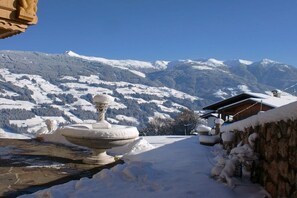 A snow-covered landscape with a stone fountain, a wooden building, and snow-covered mountains in the background. - Holiday apartment with 2 bedrooms (Fügenberg)